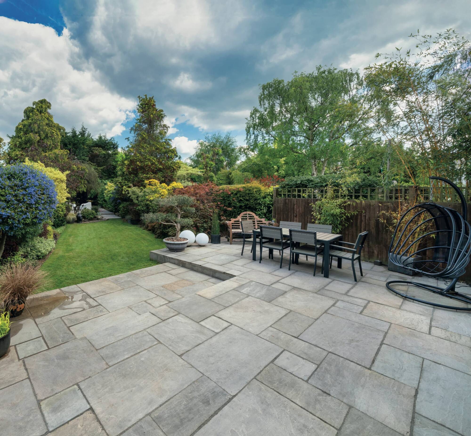 A spacious garden with a stone patio featuring a dining table and chairs. The lush greenery includes various shrubs and trees, and a neatly manicured lawn extends into the distance. The sky is partly cloudy.